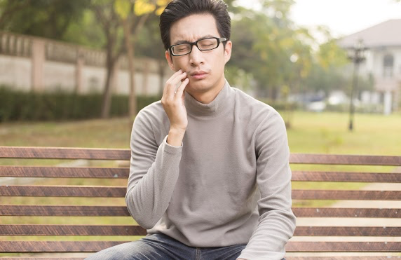 Man sitting at a park bench massaging his cheek to alleviate pain from his recent orthodontic treatment.